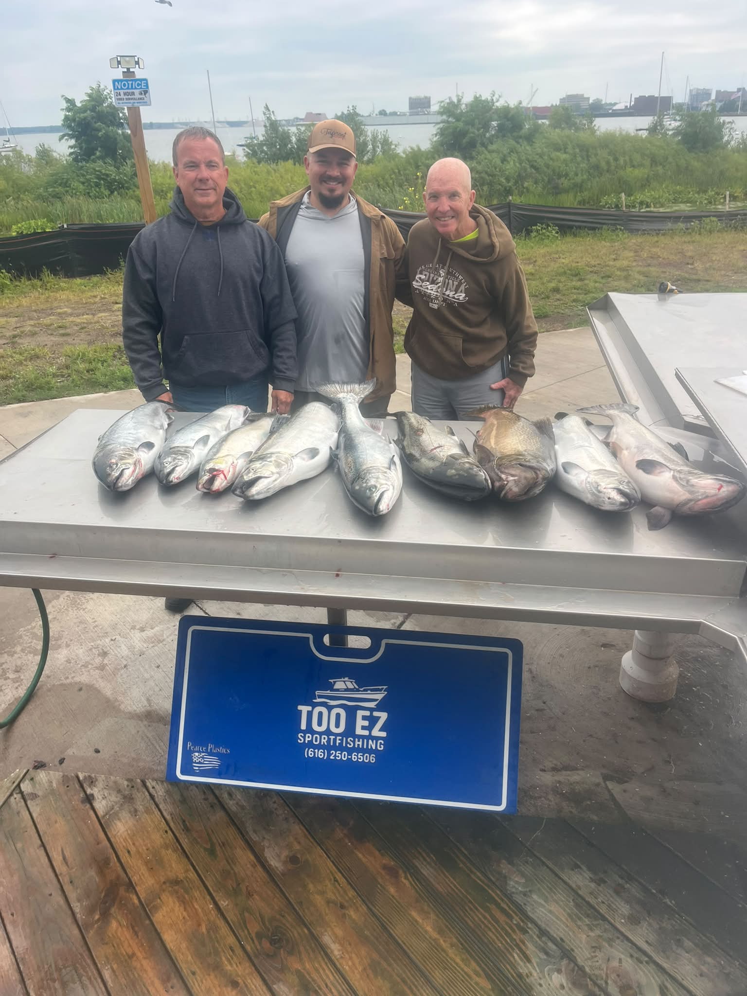 Nine salmon and trout laid out on the dock cleaning table after a morning charter out of Muskegon, with three anglers standing behind and the Too Ez Sportfishing sign in front.