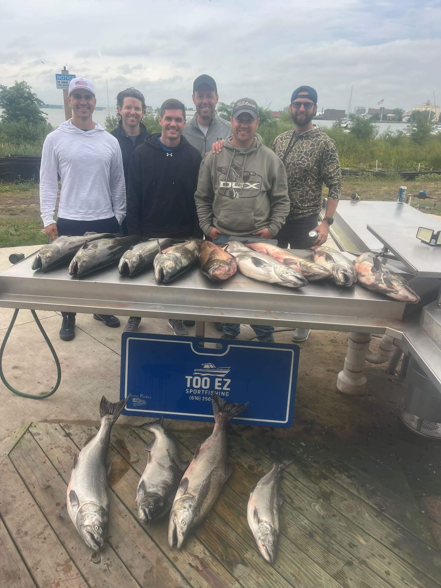 Six anglers pose with their catch of salmon and other fish displayed on a table in front of a Too Ez Sportfishing branded sign at a dock.